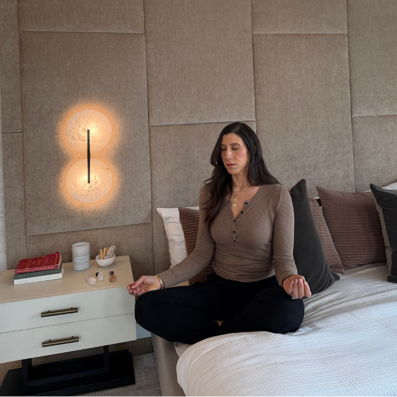 Woman meditating on a bed in a bedroom with a textured wall and bedside table.